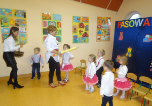 Pani dyrektor Maria Królikowska trzyma w ręku dużą żółtą kredkę, którą pasuje na przedszkolaka dzieci. Pani Ewelina stoi z koszyczkiem, podaje dyplom dla chłopca.